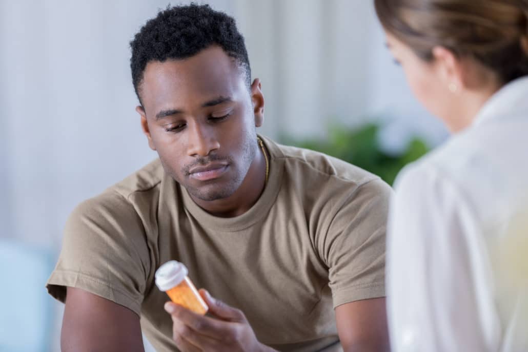 A young African American male military office sits across from his unrecognizable therapist and reads a prescription bottle.  His therapist has prescribed the medication for his depression.