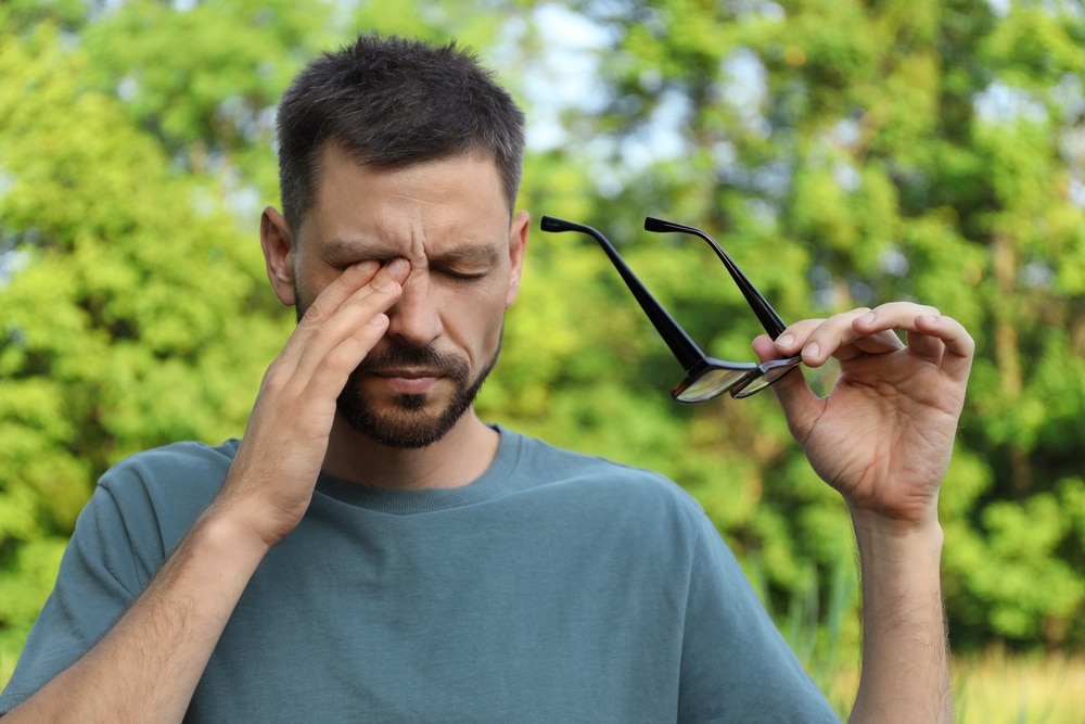 Man outdoors removes his glasses to rub his itchy eyes.